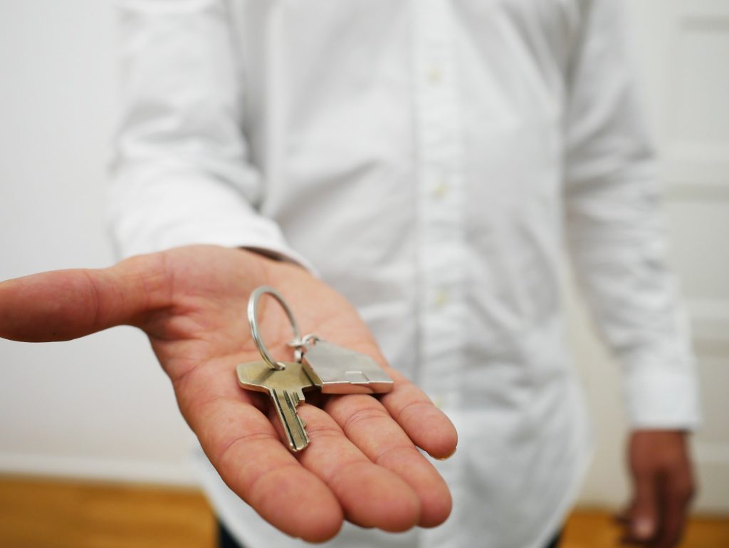 The torso of a man in a white shirt, his hand is outstretched and holding a silver key with a silver house shaped keyring.